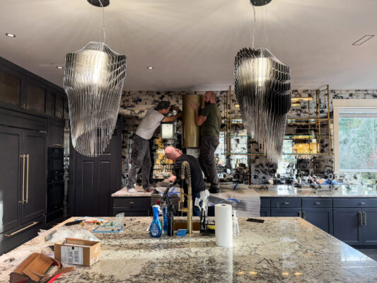 Modern kitchen renovation with workers upgrading cabinetry and features under warm chandelier lighting.